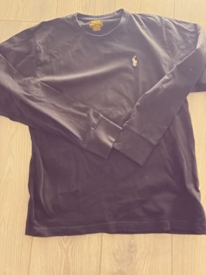 Svart långärmad tröja från Polo Ralph Lauren - Klassisk svart långärmad tröja från Polo Ralph Lauren med rund halsringning och den ikoniska broderade loggan på bröstet. Tillverkad i mjuk bomull som känns skön mot huden. Perfekt för en clean och stilren look.