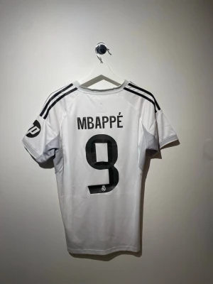 Real Madrid fotbollströja Mbappé nr 9 - Snygg vit Real Madrid matchtröja från Adidas med Mbappé och nummer 9 på ryggen. Tröjan har svarta detaljer, klubbmärke på bröstet och sponsortryck 'Emirates Fly Better' framtill. Tillverkad i lätt och funktionellt material, perfekt för fotbollsträning eller match.