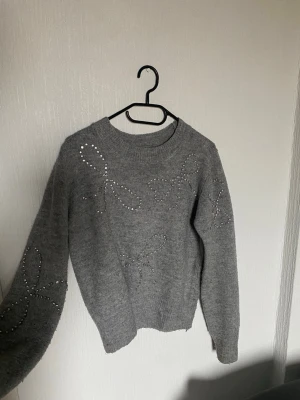 Grå stickad tröja med diamanter - Mysig grå stickad tröja med rund hals och dekorativa pärlor som bildar rosett-mönster på fram- och ärmar. En av diamanterna har trillat bort på en av diamanterna men det syns bara om man kollar noga
