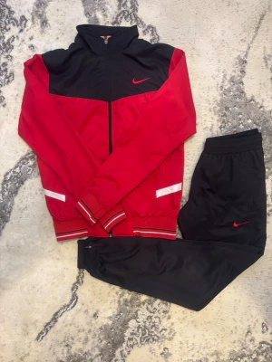 Nike röd och svart träningsset - Snyggt träningsset från Nike med röd och svart zip-jacka och matchande svarta byxor. Jackan har vita detaljer och Nike-logga på bröstet, medan byxorna har röd Nike-logga. Perfekt för gymmet eller chill. Materialet är lätt och andas.