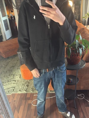 Svart hoodie från Polo Ralph Lauren - Svart hoodie med dragkedja från Polo Ralph Lauren. Klassisk design med huva, två fickor framtill och den ikoniska vita loggan broderad på bröstet. Perfekt för en avslappnad och stilren look.