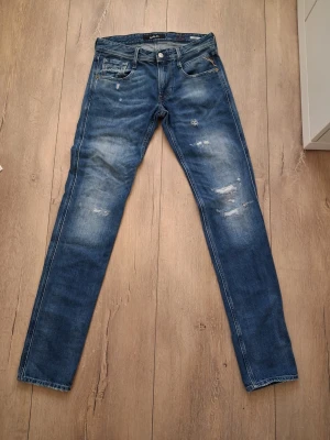 Blå replay jeans med slitningar - Säljer ett par blå slim fit jeans med snygga slitningar och tvättade detaljer på låren och knäna. Jeansen har fem fickor och klassisk passform. Perfekta för en avslappnad och trendig look.                                                                      W 28 L 32