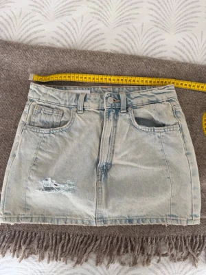 Ljusblå jeanskjol med slitning - Trendig ljusblå jeanskjol i kort modell med klassiska fem fickor och slitning framtill. Kjolen har knapp och dragkedja i midjan samt tydliga sömmar för en cool denimlook. Perfekt att matcha med sneakers eller boots.