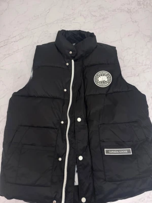 Svart dunväst från Canada Goose - Snygg svart dunväst från Canada Goose med vit dragkedja och tryckknappar. Västen har två stora fickor framtill, logotyp på bröstet och patch med märkesnamn. Insidan har ett unikt mönster i svart och vitt. Perfekt för lager-på-lager-stil.