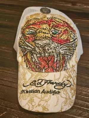 Ed Hardy keps - Snygg vit och guld Ed Hardy keps med mesh baktill och stor tiger framtill i färgglada strasstenar. Broderad logga och Christian Audigier-text på skärmen. Kepsen har detaljer i glitter och ett metallmärke på toppen. Perfekt statement piece för dig som gillar streetwear.