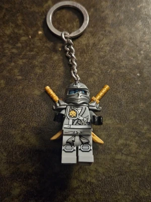 Nyckelring med LEGO Ninjago-figur - Cool nyckelring med en grå LEGO Ninjago-ninjafigur. Figuren har detaljerad rustning, blå ögon och två gyllene svärd på ryggen. Perfekt accessoar för dig som gillar LEGO eller Ninjago och vill ha något unikt till dina nycklar.