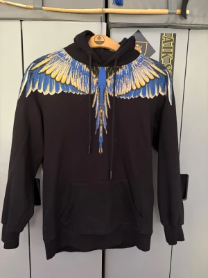 Svart hoodie med färgglada vingar - Svart hoodie från Marcelo Burlon med ikoniskt vingtryck i blått, gult och vitt över axlar och bröst. Hoodien har huva med dragsko och en stor magficka. Tillverkad i mjuk bomull, perfekt för en cool streetwear-look. M passar L