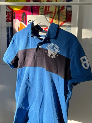 Blå pikétröja från Peak Performance - Snygg blå pikétröja från Peak Performance med svart horisontell rand och vit broderad logga på bröstet. Tröjan har klassisk krage, knappar vid halsen och tryckt nummer på ärmen. Perfekt för en sportig och avslappnad stil.