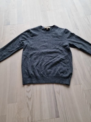 Mörkgrå rundhalsad kashmirtröja från Burberry  - En stilren mörkgrå tröja med rund halsringning. Tröjan är tillverkad i mjuk ull och har långa ärmar samt ribbade muddar vid ärmslut och nederkant. Perfekt för lager-på-lager och enkel att matcha med olika outfits.