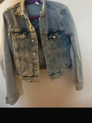 Ljusblå klassisk jeansjacka - Snygg ljusblå jeansjacka med klassisk krage, två bröstfickor med lock och knappar framtill. Jackan har raka ärmar och en något kortare passform. Perfekt att slänga över en hoodie eller t-shirt för en avslappnad look.