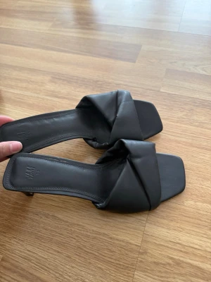 Svarta klackar från H&M - Stilrena svarta klackar med en liten klack från H&M med breda, vadderade remmar i korsad design. Sandalerna har fyrkantig tå och låg klack, tillverkade i konstläder för en trendig look. Perfekta för dig som gillar minimalistisk och modern stil.