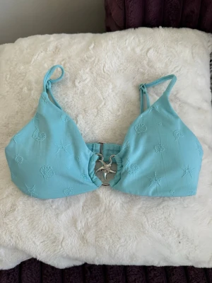 Turkos bikiniöverdel med snäckdetaljer - Snygg turkos bikiniöverdel med broderade snäckor och sjöstjärnor. Toppen har smala axelband och en unik silverfärgad snäcka i metall framtill. Perfekt för strandhäng och sommarvibbar! Obs katt finns i hem!