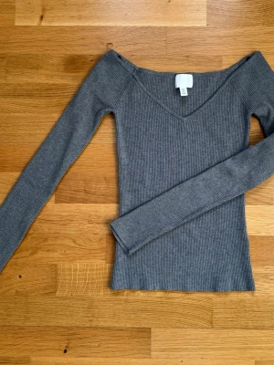 Grå ribbad långärmad topp H&M - Snygg grå ribbad topp från H&M med v-ringning och lång ärm. Modellen är figurnära och har en stilren look som passar perfekt till jeans eller kjol. Materialet är mjukt och stretchigt för en bekväm känsla.