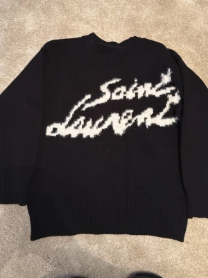 Svart Saint Laurent stickad tröja - Cool svart stickad tröja från Saint Laurent med vit, fuzzy logotyp i grafisk stil över bröstet. Tröjan har rund halsringning och ribbstickade muddar vid ärmslut och nederkant. Tröjan är tjock, varm samt känns väldigt skön att ha på sig. Väldigt bra kvalitet. Perfekt för dig som gillar streetstyle och designerplagg. 