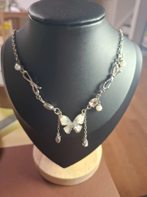 Silverfärgat halsband med fjäril och pärlor - Ett snyggt silverfärgat halsband med en glittrig fjäril i mitten och små pärlor längs kedjan. Kedjan har dekorativa detaljer och två hängande kedjor med pärlor. Perfekt accessoar för att lyfta din stil.