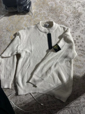 Vit stickad tröja från Stone Island - Säljer en stilren vit stickad tröja från Stone Island med klassisk patch på ärmen. Tröjan har ribbade muddar, hög krage och är gjord i ett mjukt stickat material. Perfekt för dig som gillar enkel men snygg design.