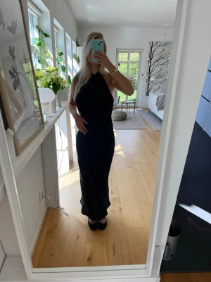 Svart långklänning med halterneck - Stilren svart långklänning med halterneck och ärmlös design. Klänningen har en elegant och figurnära passform som ger en snygg siluett. Klänningen är glansig och djup i ryggen😊