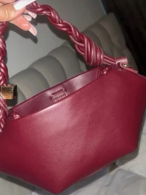 Vinröd handväska med flätat handtag - Snygg vinröd handväska i slätt läder med ett unikt flätat handtag. Väskan har en modern, kantig form och detaljer i guld. Perfekt accessoar för att lyfta din outfit. Köpt från Plick innan 💖