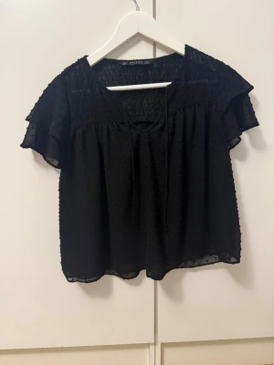 Svart blus från Zara Basic - Snygg svart blus från Zara Basic med korta, volangiga ärmar och diskret struktur i tyget. Blusen har en lös passform och knytband vid halsen. Perfekt att styla med jeans eller kjol för en trendig look.