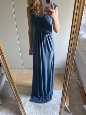 Blå långklänning med v-ringning - Elegant blå långklänning med djup v-ringning och draperad överdel. Klänningen har breda axelband och ett mjukt, fallande tyg som ger en snygg siluett. Perfekt för speciella tillfällen som exempelvis bal eller bröllops gäst.