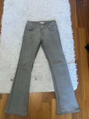 Grå bootcut jeans från Gina tricot - Snygga grå jeans från Gina tricot med bootcut passform och klassisk femficksdesign. Jeansen har dubbla knappar i midjan och bälteshällor. Perfekta för dig som gillar en avslappnad men trendig stil.