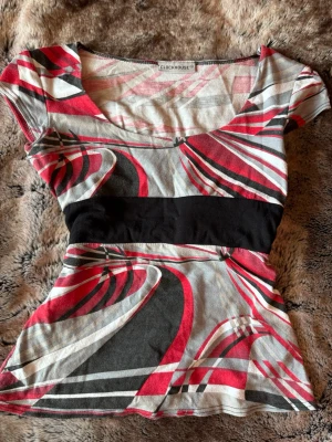 Mönstrad topp från Clockhouse - Snygg vintage topp från Clockhouse med abstrakt mönster i rött, svart, grått och vitt. Modellen har korta ärmar, markerad midja med svart band och rund halsringning. Perfekt för dig som gillar färg och grafiska prints.