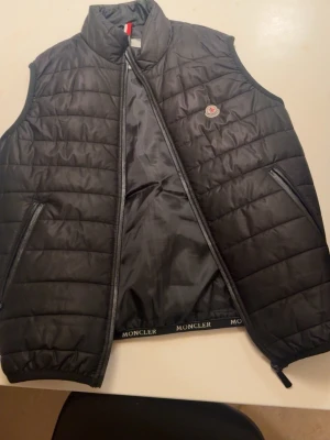 Svart dunväst från Moncler - Snygg svart dunväst från Moncler med quiltad design och hög krage. Västen har dragkedja framtill, två fickor med dragkedja och Moncler-logga på bröstet. Perfekt för lager-på-lager och streetstyle. Materialet är glansigt och lätt, med klassisk Moncler-detalj på insidan. Storlek xs 