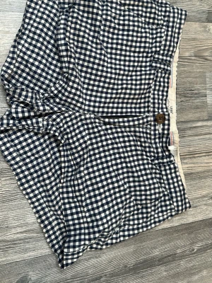 Rutiga blåvita shorts med knappar - Snygga shorts med blåvitt rutmönster och två bakfickor med bruna knappar. Klassisk passform och mjukt, tunt tyg som känns skönt på sommaren. Perfekta för dig som gillar en avslappnad men ändå stilren look.