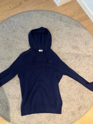 Mörkblå hoodie från Bvaldi - Mysig mörkblå hoodie från Bvaldi med dragsko i huvan och diskret broderad logga på bröstet. Tillverkad i mjukt stickat material som känns skönt mot huden. Perfekt för dig som gillar en enkel och stilren look.