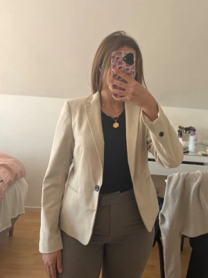 Beige figursydd kavaj - Stilren beige kavaj med figurnära passform och klassisk krage. Kavajen har en knapp framtill och dekorativa knappar vid ärmsluten. Perfekt att styla med både jeans och kostymbyxor för en snygg look.