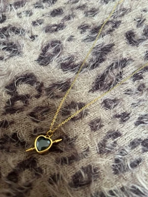 Halsband med svart hjärthänge guld spjut - Guldigt halsband med ett hjärtformat svart hänge och en dekorativ tvärgående detalj. Kedjan är tunn och elegant, och hänget har en glansig finish. Perfekt accessoar för att lyfta din outfit med en cool och trendig touch. Rostfri