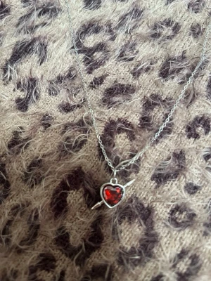 Halsband med röd hjärtberlock spjut silver - Snyggt halsband i silverfärg med en hjärtformad berlock i rött glas eller sten. Hjärtat är infattat i metall och har en pil genom sig, vilket ger en cool och lekfull vibe. Perfekt accessoar för att lyfta din outfit. Rostfri 