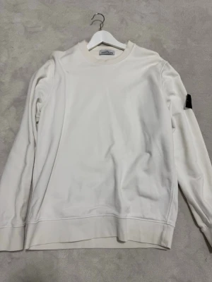 Vit sweatshirt från Stone Island - Säljer en stilren vit sweatshirt från Stone Island med klassisk rund hals och den ikoniska loggan på vänster ärm. Tröjan är i mjukt material och har ribbade muddar vid ärmslut och nederkant. Perfekt för en clean och trendig look.