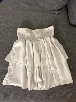 Vit volangkjol med smock i midjan - Supersöt vit kjol med bred smock i midjan och dubbla volanger. Kjolen är kort och har en flowy känsla med glansigt tyg som ger en fräsch look. Perfekt för att styla med en crop top eller stickad tröja. Osäker på storlek men skulle vilja säga xs💕