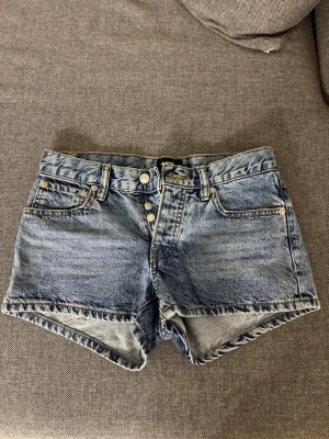 Blå denimshorts från lager 157 - Klassiska blå denimshorts från lager 157 med hög midja och knappgylf. Shortsen har fem fickor och en avslappnad passform, perfekta för varma dagar. Snygg tvättad look och slitna detaljer ger en cool vibe.