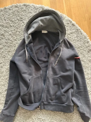 Mörkgrå hoodie från Moncler - Snygg mörkgrå hoodie från Moncler med dragkedja, fickor framtill och huva med grå insida. Ärmen har Moncler-logga och en röd, vit och blå rand. Perfekt för en avslappnad och trendig stil. 