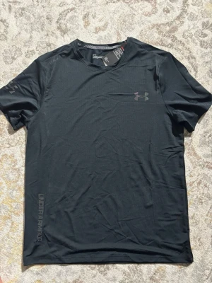 Svart tränings-tshirt från Under Armour - Svart t-shirt från Under Armour med diskret logga på bröstet och tryck längs sidan. Tillverkad i ett lätt och snabbtorkande funktionsmaterial, perfekt för träning och sport. Klassisk rund hals och korta ärmar för maximal rörelsefrihet.