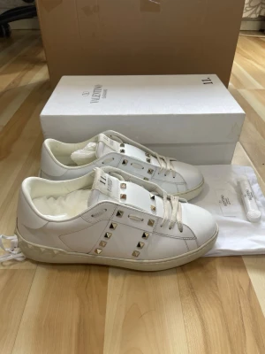 Vita Valentino Rockstud sneakers - Snygga vita sneakers från Valentino med ikoniska Rockstud-nitar i silver längs sidorna och på plösen. Skorna är låga och har en klassisk siluett med vita skosnören och detaljer i läder. Perfekta för dig som gillar exklusiv och trendig streetstyle.
