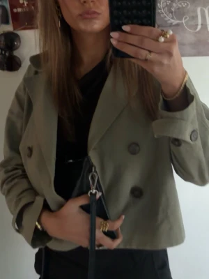 Kort grön trenchcoat med dubbelknäppning - Snygg kort trenchcoat i olivgrönt med klassisk dubbelknäppning och bred krage. Jackan har en rak passform och är tillverkad i ett lätt tyg, perfekt för lager-på-lager. Inga synliga loggor eller märken. Passar dig som gillar stilrena och trendiga ytterplagg.