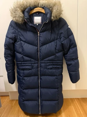 Mörkblå lång dunjacka Tommy Hilfiger - Snygg mörkblå lång dunjacka från Tommy Hilfiger med fuskpäls på huvan och dragkedja framtill. Jackan har quiltad design, två fickor och en hög krage. Perfekt för kalla vinterdagar och riktigt varm.