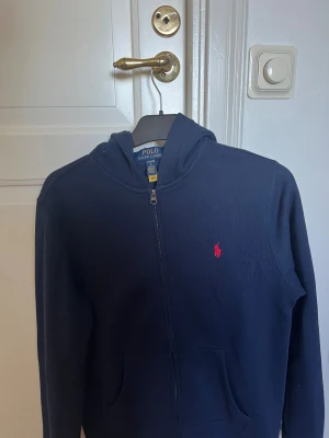 Mörkblå hoodie från Polo Ralph Lauren - Snygg mörkblå hoodie med dragkedja från polo Ralph lauren. Tröjan är helt ny, sälj pga att den är för liten 