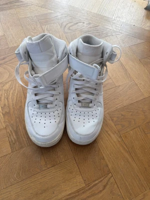 Nike Air Force 1 vita high sneakers - Klassiska vita Nike Air Force 1 high sneakers med högt skaft och kardborreband runt ankeln. Skorna har perforerad tå för ventilation, tjock sula och ikonisk Nike-logga på hälen. Tillverkade i läder med textilfoder och snörning framtill.