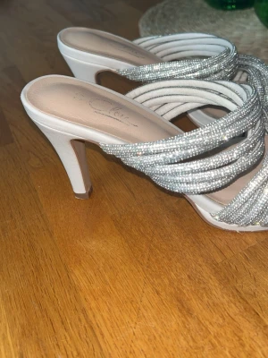 En festsko beige glittriga  - Säljer ett par snygga pumps med öppen tå och höga klackar. Skorna är vita med flera glittriga silverremmar över foten som ger en riktigt festlig look. Perfekta för dig som vill sticka ut och glänsa lite extra. Märket är okänt men designen är supertrendig.