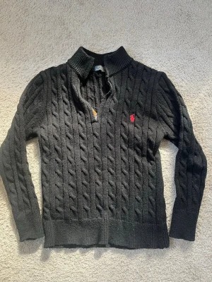  Polo Ralph Lauren - Svart kabelstickad tröja från Polo Ralph Lauren med röd broderad logga på bröstet. Tröjan har v-ringning, ribbstickad krage och långa ärmar. Perfekt för dig som gillar klassisk och stilren design.