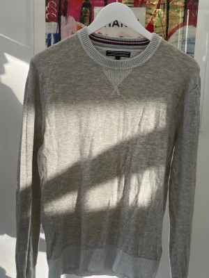 Ljusgrå tröja Tommy Hilfiger - Ljusgrå långärmad tröja från Tommy Hilfiger med ribbade muddar och krage. Rund halsringning med snygg detalj i vitt och röd-svart rand på insidan av kragen. Perfekt för en stilren och avslappnad look. Passar nog även M.