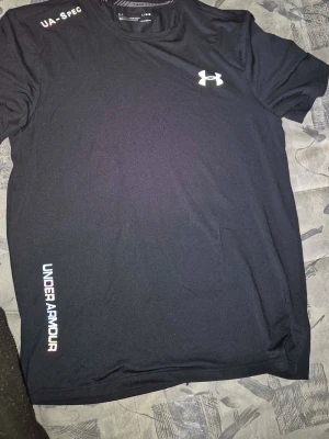 Svart träningströja från Under Armour - Svart sportig t-shirt från Under Armour med logga på bröstet och texten UNDER ARMOUR längs sidan. Tillverkad i ett lätt och snabbtorkande material som passar perfekt för träning och aktivitet. Klassisk rund hals och kort ärm för maximal rörelsefrihet.