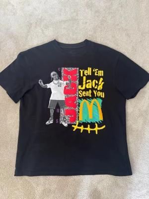 Svart Travis Scott x McDonald's T-shirt - Cool svart T-shirt med färgglatt tryck framtill med texten 'Tell 'Em Jack Sent You' och McDonald's-logga. På baksidan finns ett blått grafiskt tryck. Perfekt för dig som gillar streetwear och unika samarbeten. Tröjan är i storlek M