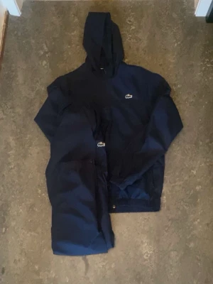 Mörkblå vindjacka från Lacoste - Snygg mörkblå vindjacka från Lacoste med huva och dragkedja. Jackan har den klassiska krokodilloggan på bröstet och framtill. Perfekt för blåsiga dagar och enkel att matcha med olika outfits.