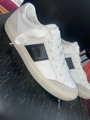Vita Lacoste sneakers med svart detalj - Snygga vita sneakers från Lacoste med svart panel och krokodillogga på sidan. Skorna har vita tjocka snören, perforerad tå och beige detaljer vid tån och hälen. Tillverkade i läder och mocka för en clean och lyxig look. Storlek 44.