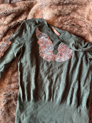 Grön stickad tröja med spetsdetaljer - Mörkgrön stickad tröja från Saint Tropez med unika spetsdetaljer i vitt och rosa vid halsringningen och på ena ärmen. Tröjan har ribbstickad nederkant och ärmar, ryggen har vingar i silver 🪽 